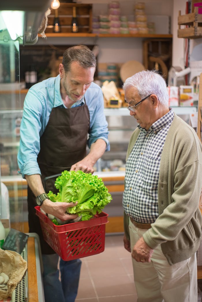 Elderly man shopping at a local market assisted by a grocery clerk holding fresh lettuce.